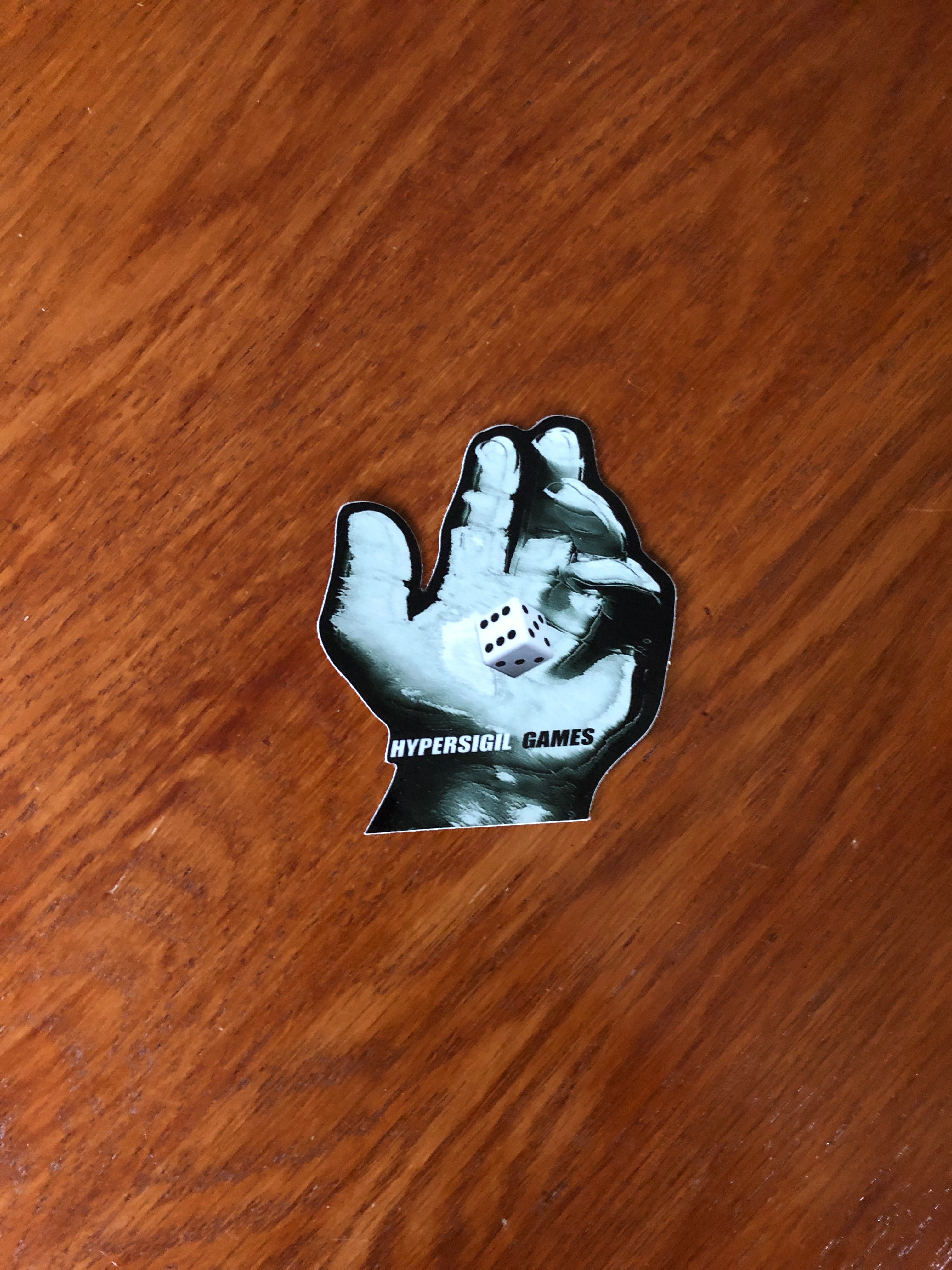 a sticker that portrays a hand with a dice in the middle as if in motion with text reading "hyper sigil games"